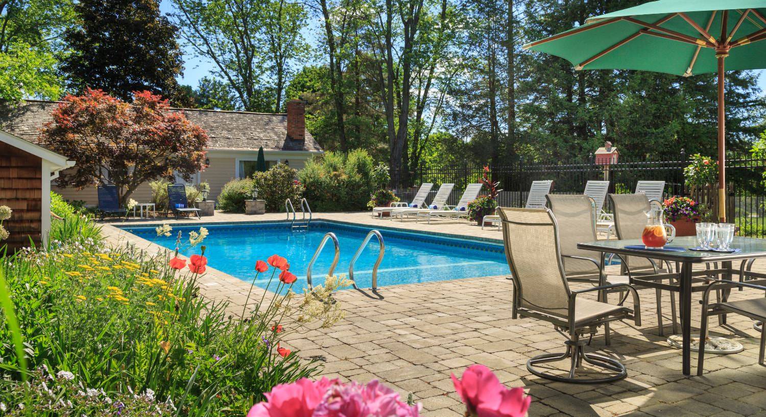 Pool area surrounded by brick patio with patio table and chairs, green umbrella, black wrought-iron fencing, large treen trees, and a lot of bushes and shrubs