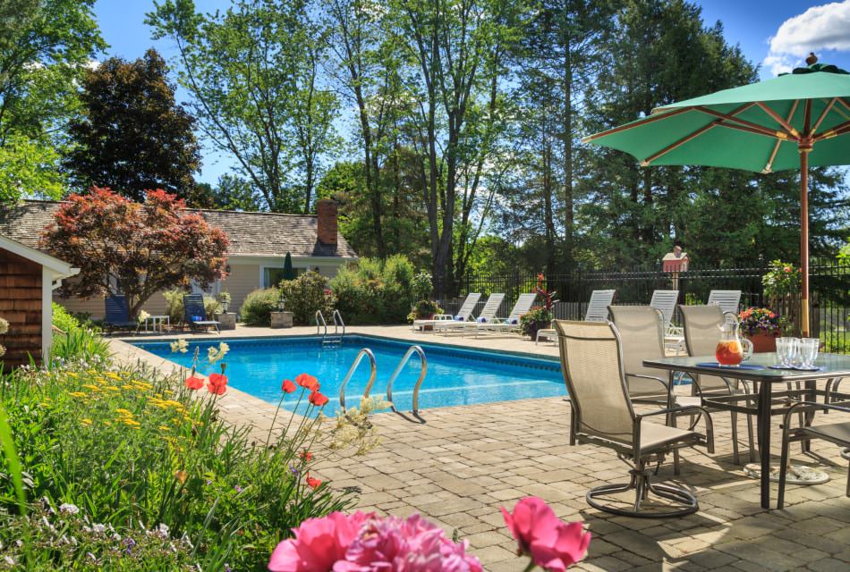 Exterior view of the property's pool surrounded by a stone brick patio, pool furniture, patio table with chairs and umbrella, and green trees, shrubs, and vegetation