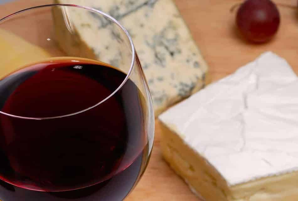 Close up view of a glass of red wine, two types of cheese and one red grape on wooden board