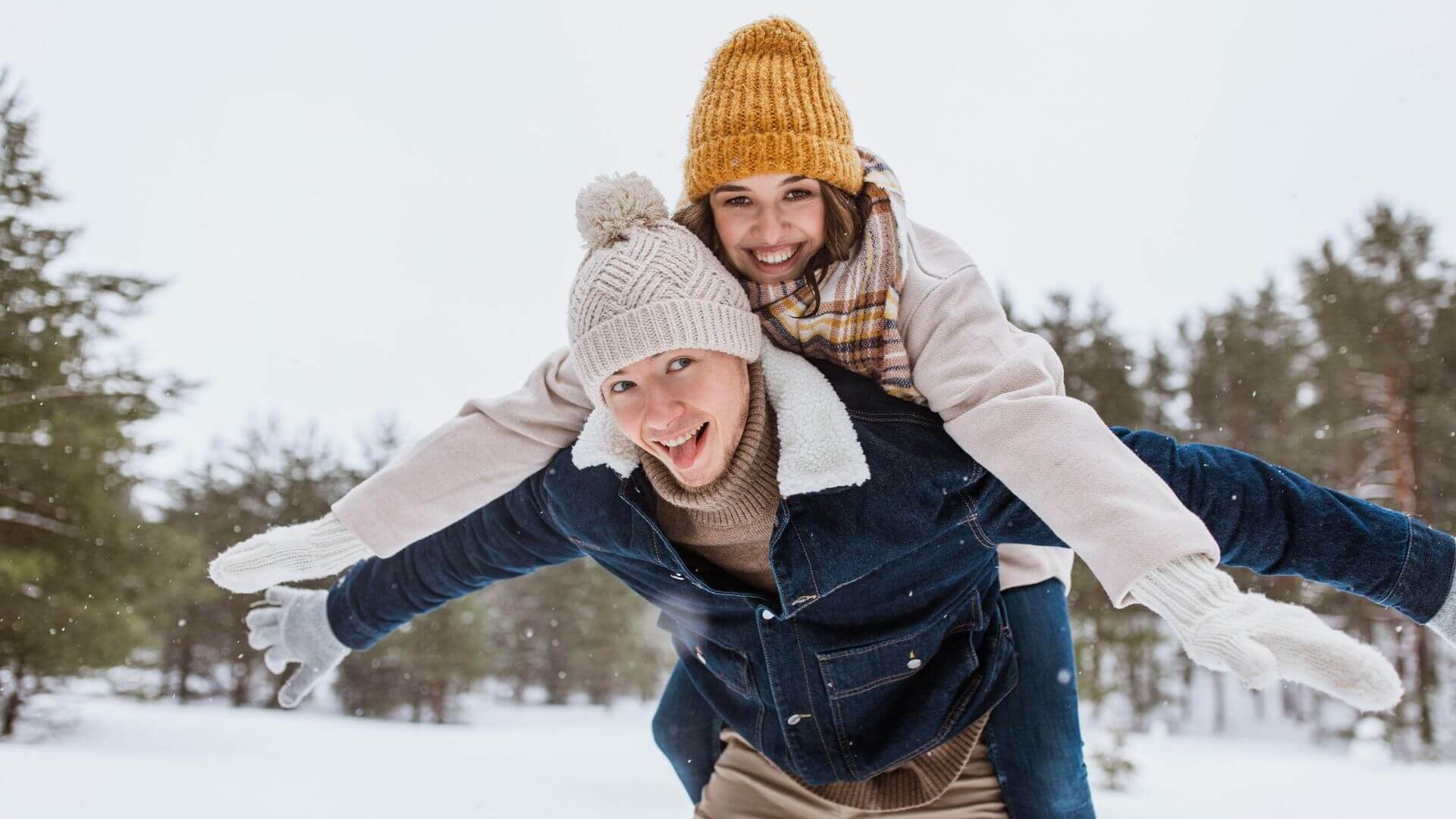 a smiling couple dressed in winter clothes plays in the snow with trees in the background