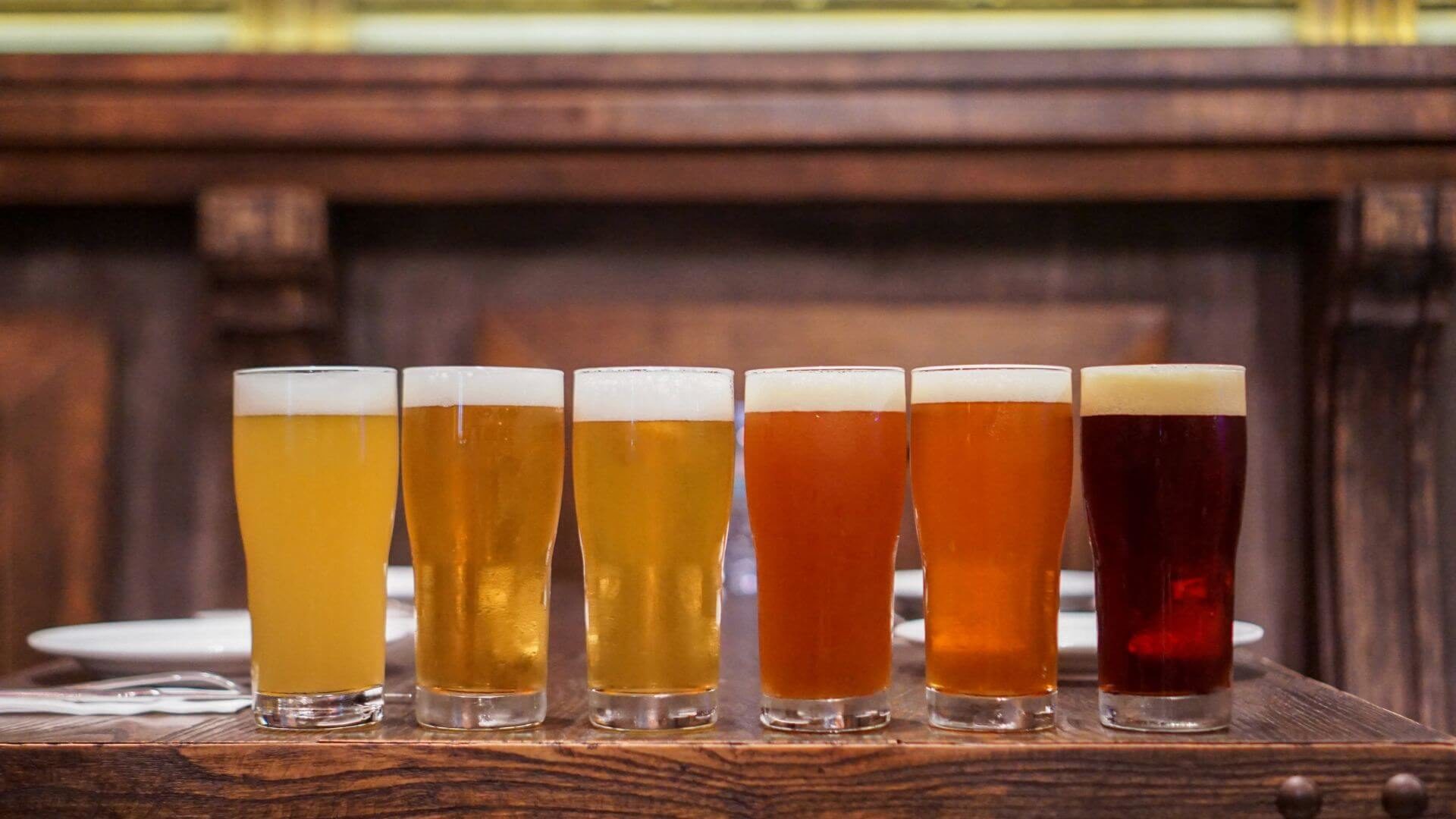 Six clear glasses sitting on wooden table with various varieties of beer with foam on top.