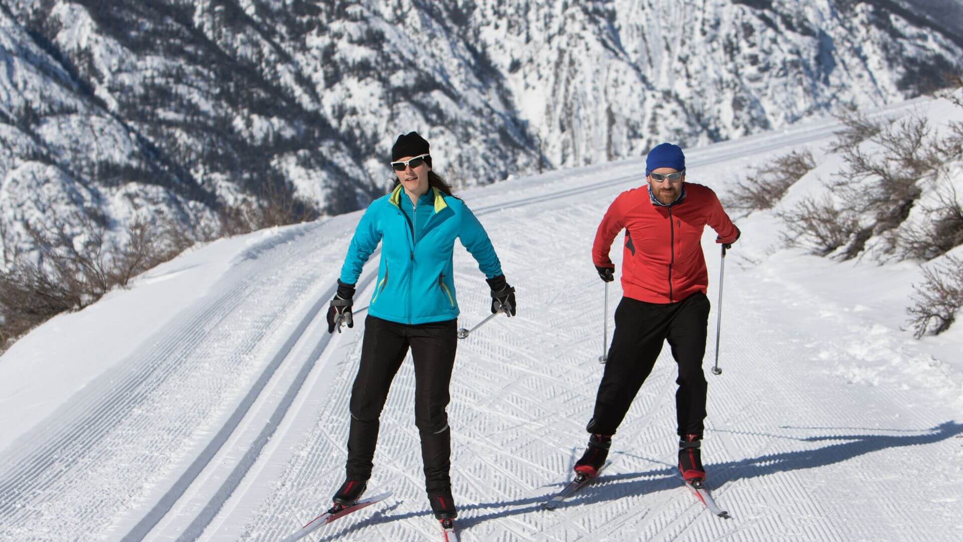 Man in red jacket and woman in blue jacket cross country skiing along a well traveled trail.