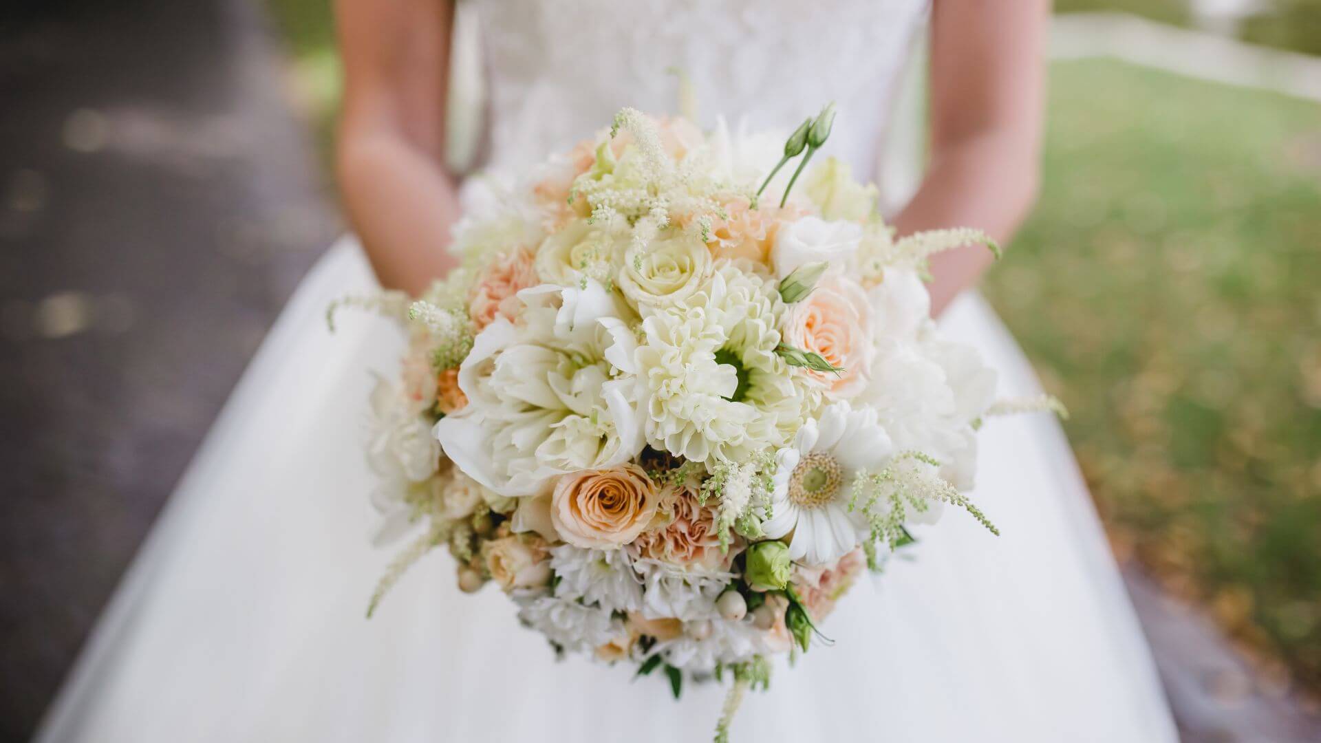 Bride in white dress standing while holding a bouquet of peach roses and other white flowers.
