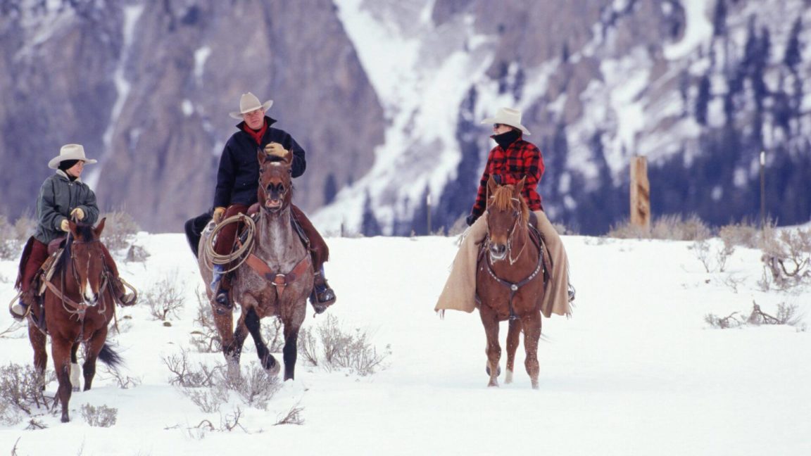Group of people wearing cowboy hats and wearing winter coats on brown horses riding through snowy terrain.