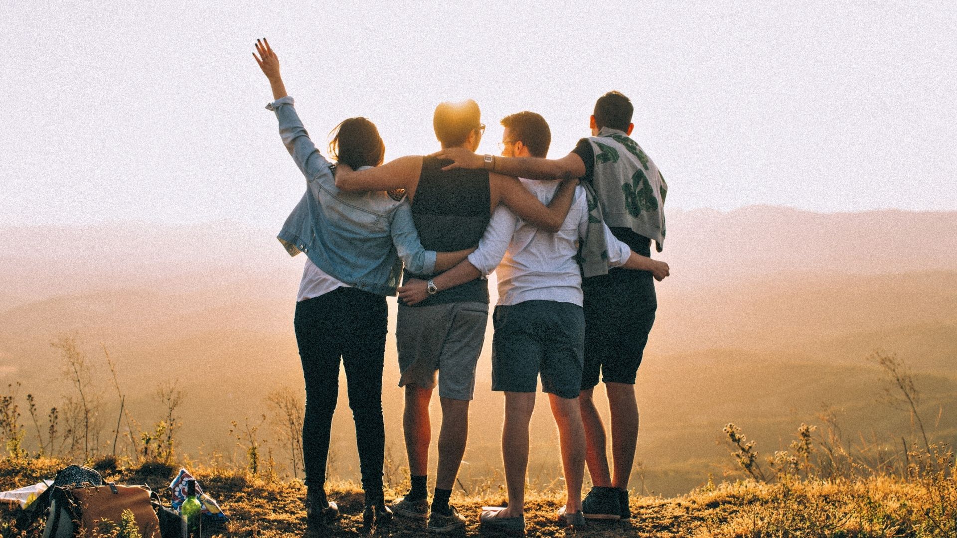 group of friends with their arms around each other standing together on a hilltop watching the sunset