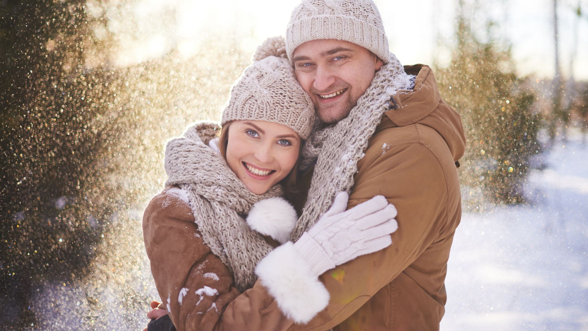 A couple wrapped in each other's arms dressed in warm winter clothes with the sun behind them as they stand outside in the snow.