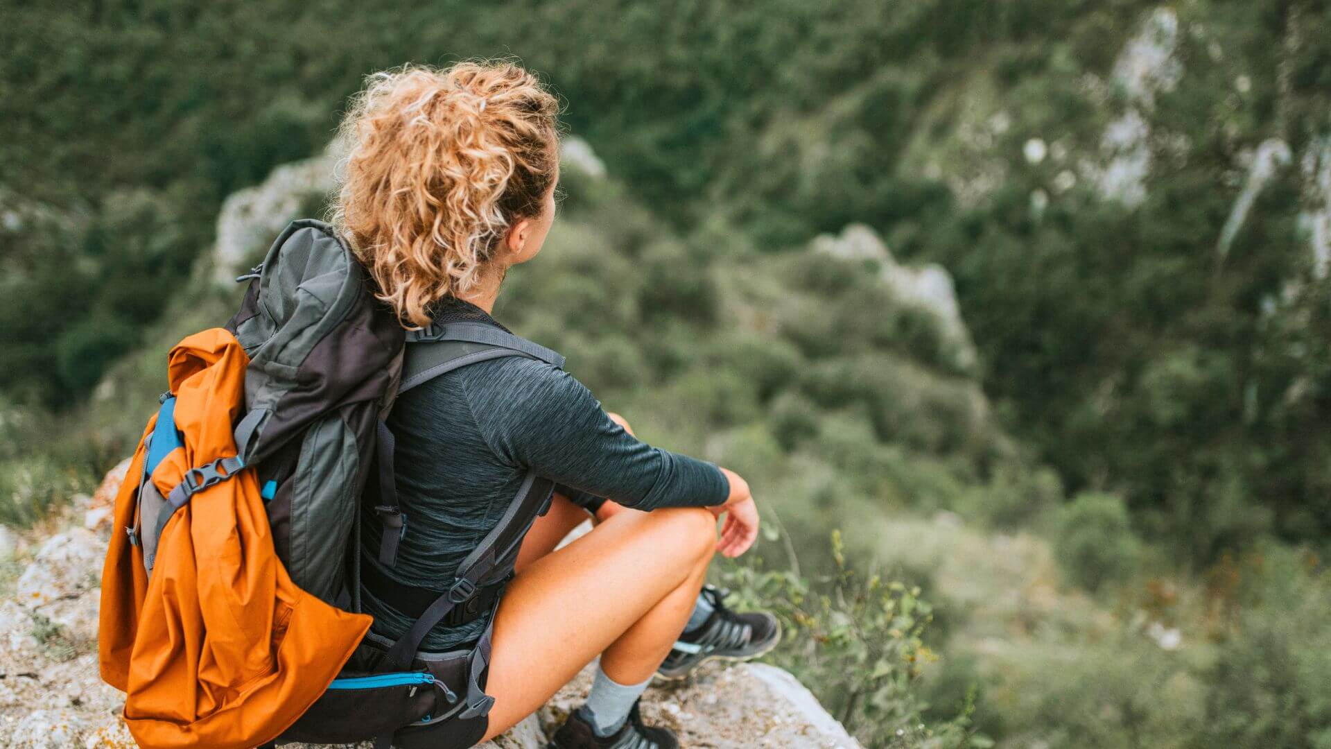 A hiker with a large backpack sits on a rocky overlook, facing a forested landscape.