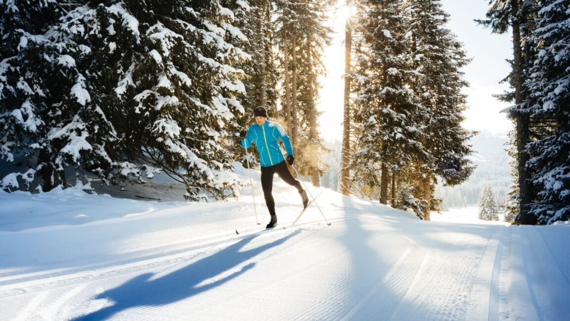 A man is cross-country skiing with the winter sun shining through the trees in the background.