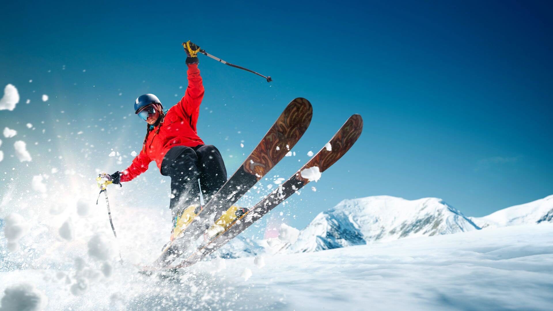 Man wearing red jacket, goggles and helmet downhill skiing with snow flying