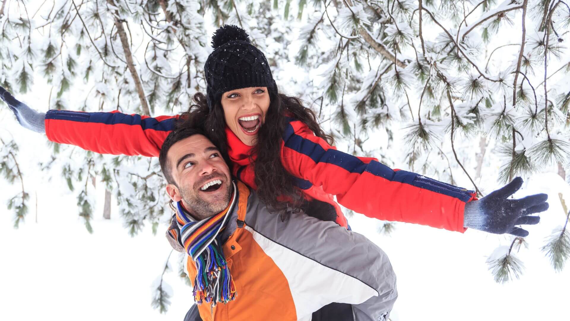Smiling woman in red and blue coat riding piggy back on smiling man in orange, gray and white coat standing in snow under evergreen trees