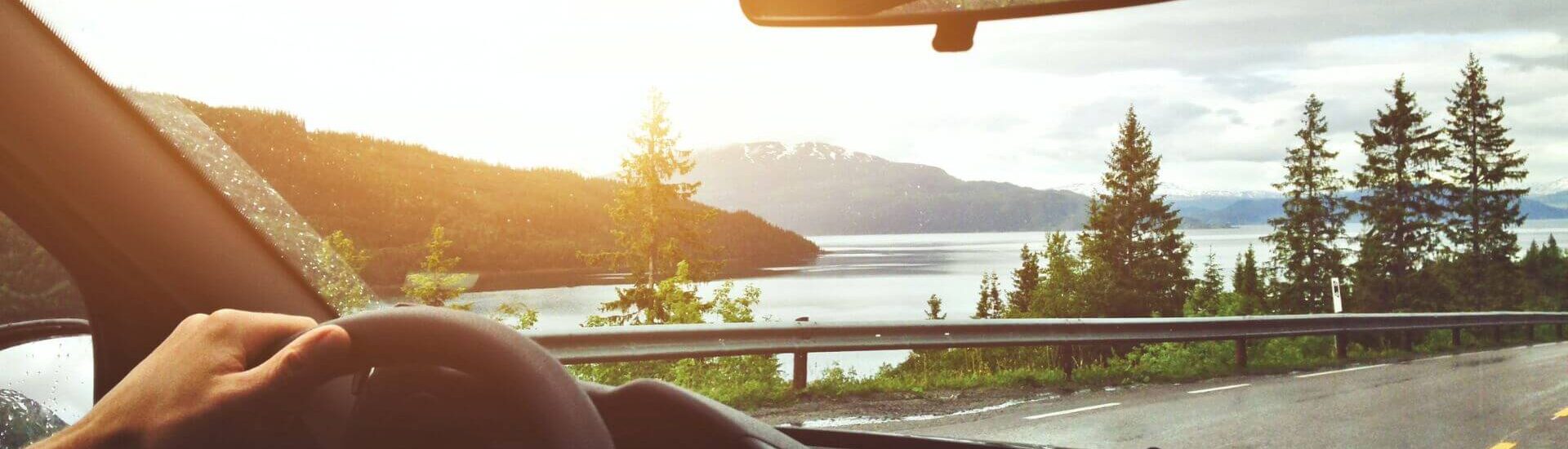 View from inside a car driving along a scenic road with a lake, mountains, and trees at sunset.