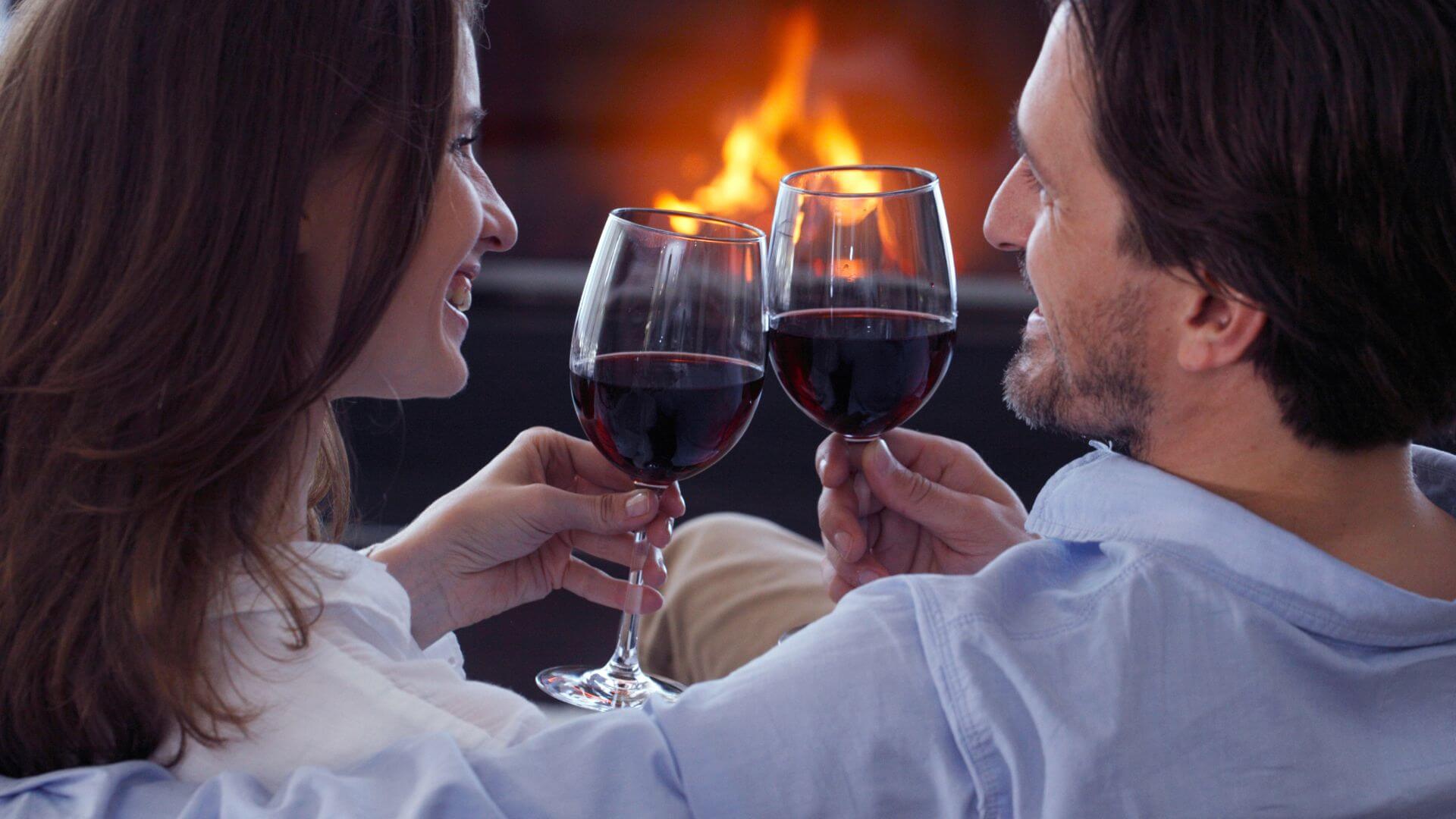 Smiling couple sitting in front of burning fireplace clinking red wine glasses together.
