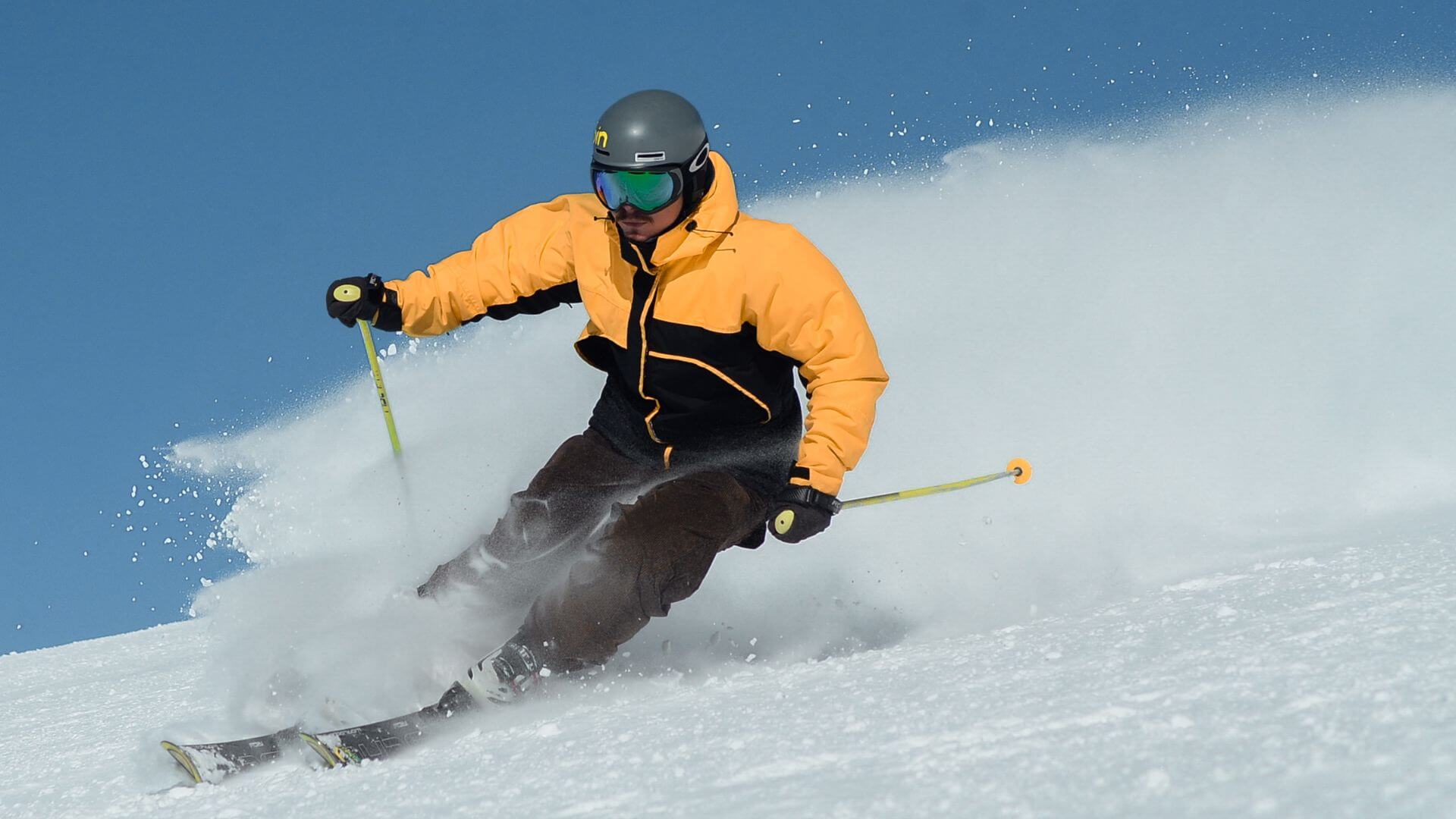 Man in Gold and Black jacket wearing gray helmet and goggles skiing downhill with snow blowing.