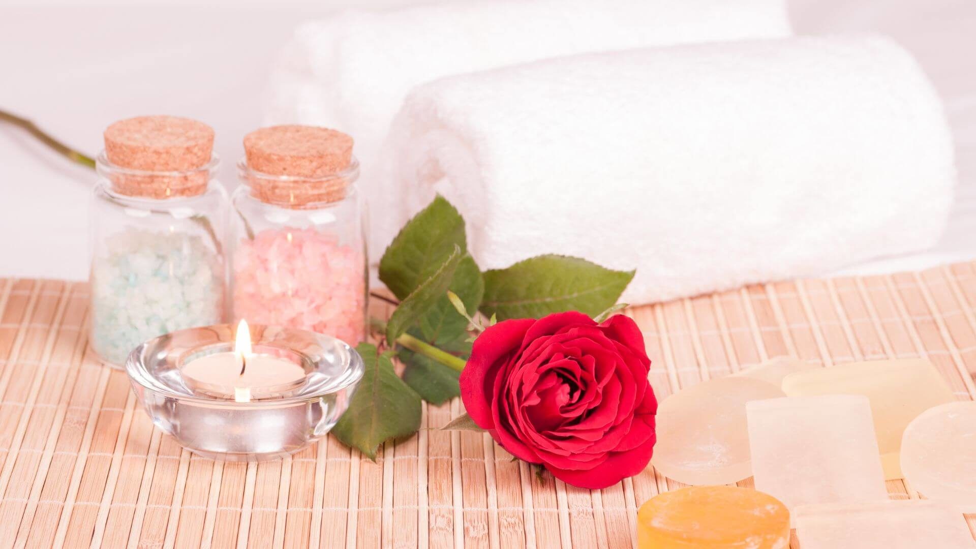 Spa table holding glass jars with bath salts, lit candle, rolled town and red rose.