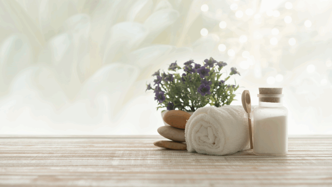 spa setting with a rolled towel, jar of bath salts, smooth stones, and flowers on a wooden surface
