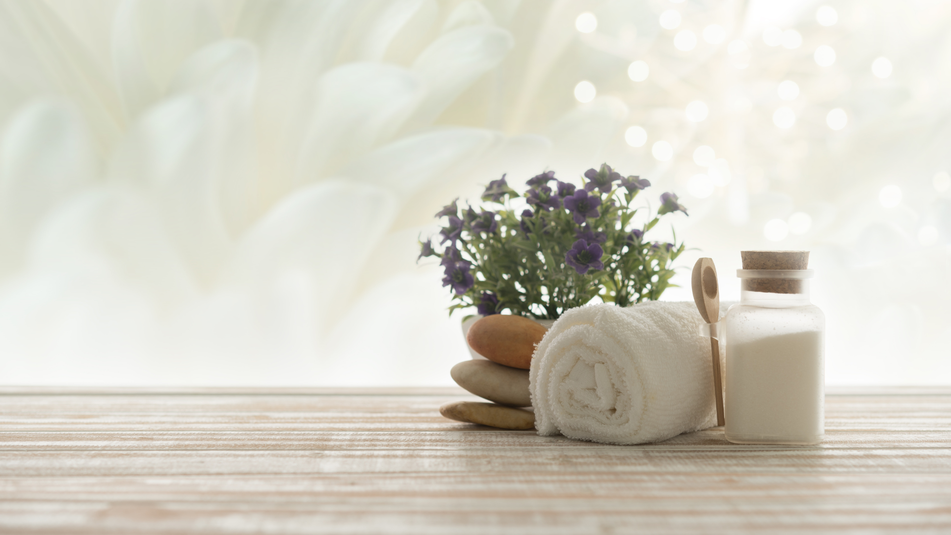 spa setting with a rolled towel, jar of bath salts, smooth stones, and flowers on a wooden surface