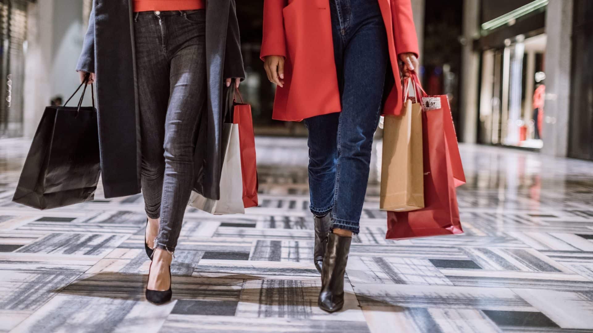 View of two people waist down holding shopping bags and walking through a mall.