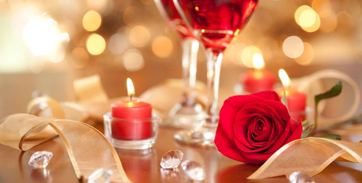 Sparkling light background with candles, a red rose, twirling ribbon and two glasses of wine