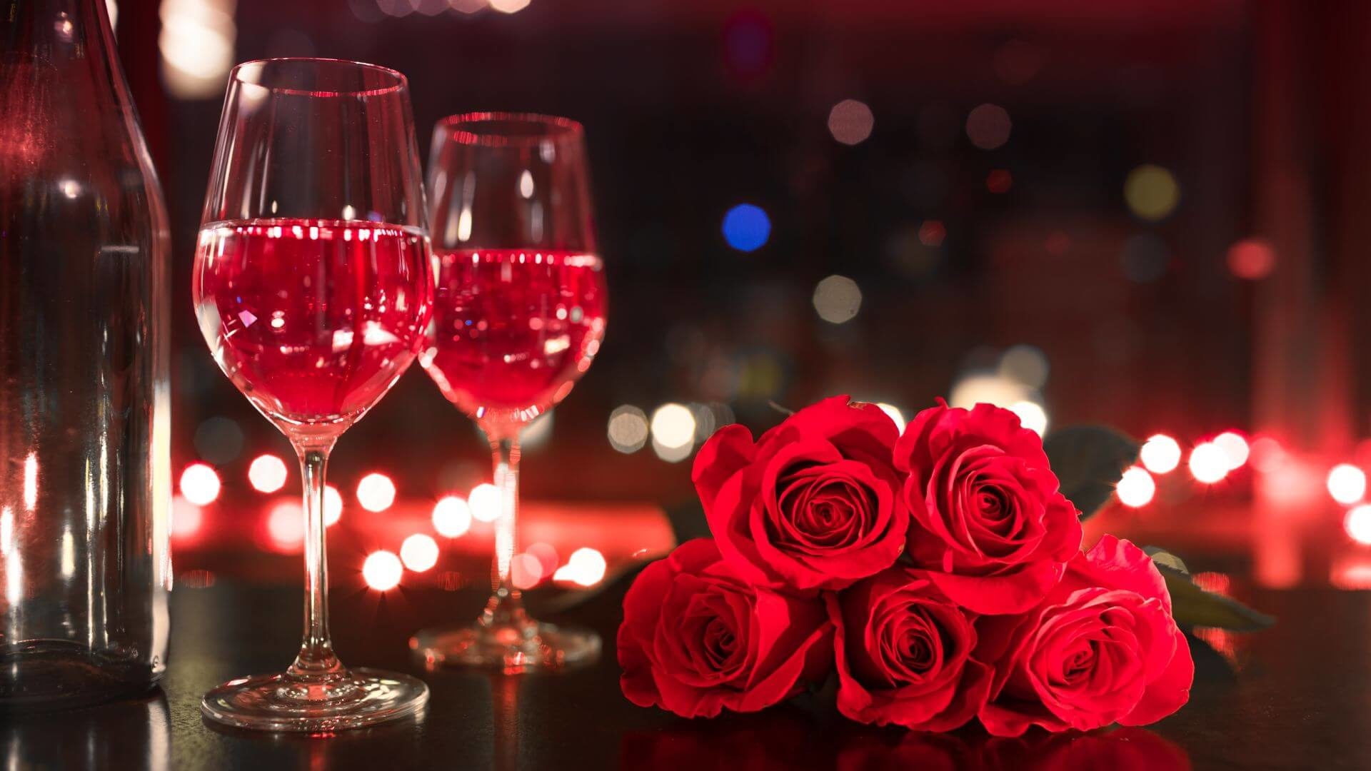 Filled wine glasses by bottle with red roses laying on table in dimly lit room.