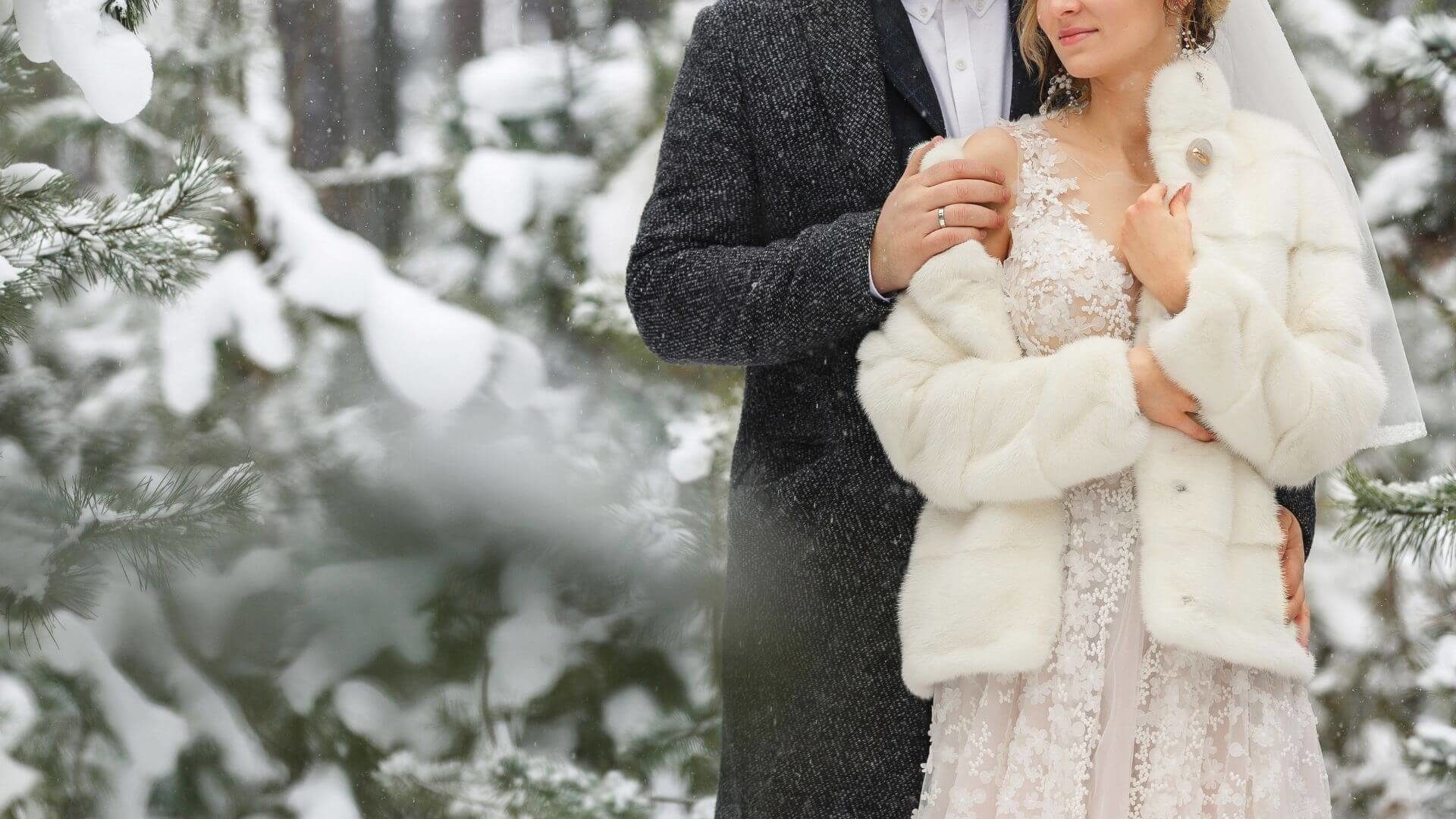 A groom with his arms wrapped around his bride wearing fur coat in a forest of snow-covered firs.