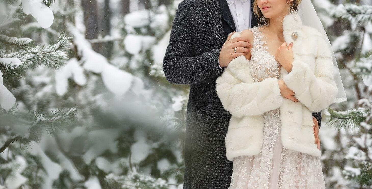 A groom with his arms wrapped around his bride in a forest of snow-covered firs.