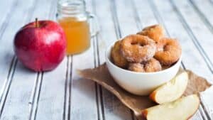 A bowl of apple cider donuts with a fresh apple and apple cider on the side