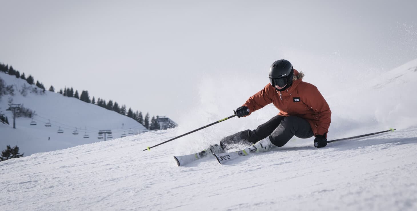 man with helmet wearing red jacket downhill skiing with snow flying up behind ski lift in distance