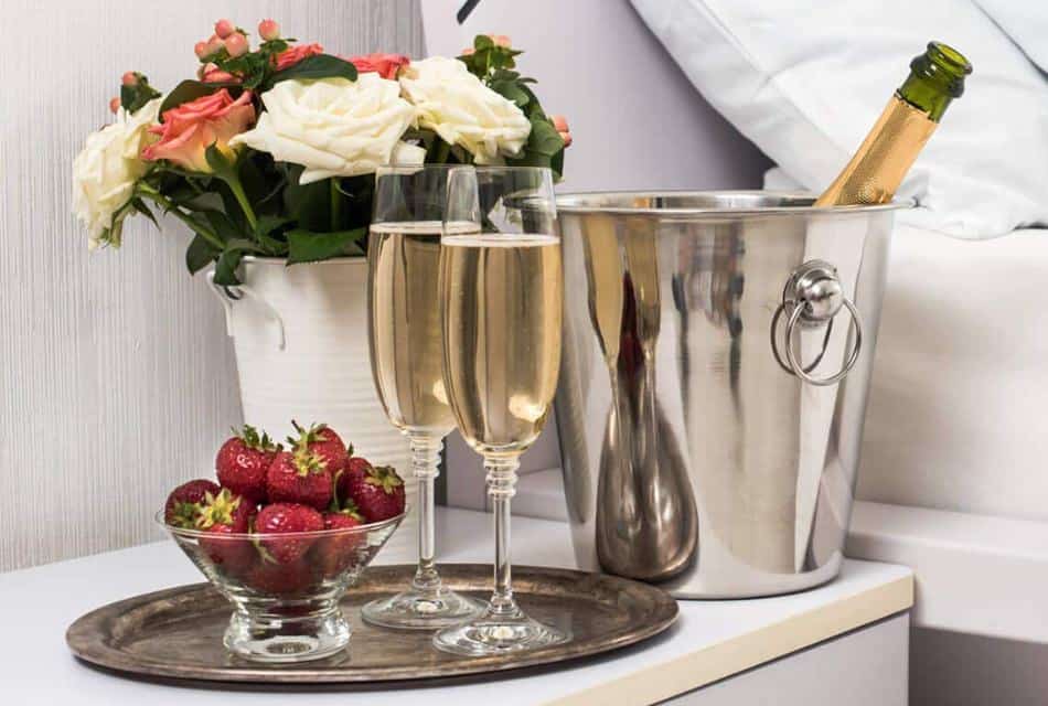 Bottle of champagne chilling in a silver tub, bowl of strawberries, vase with flowers, and flutes filled with champagne