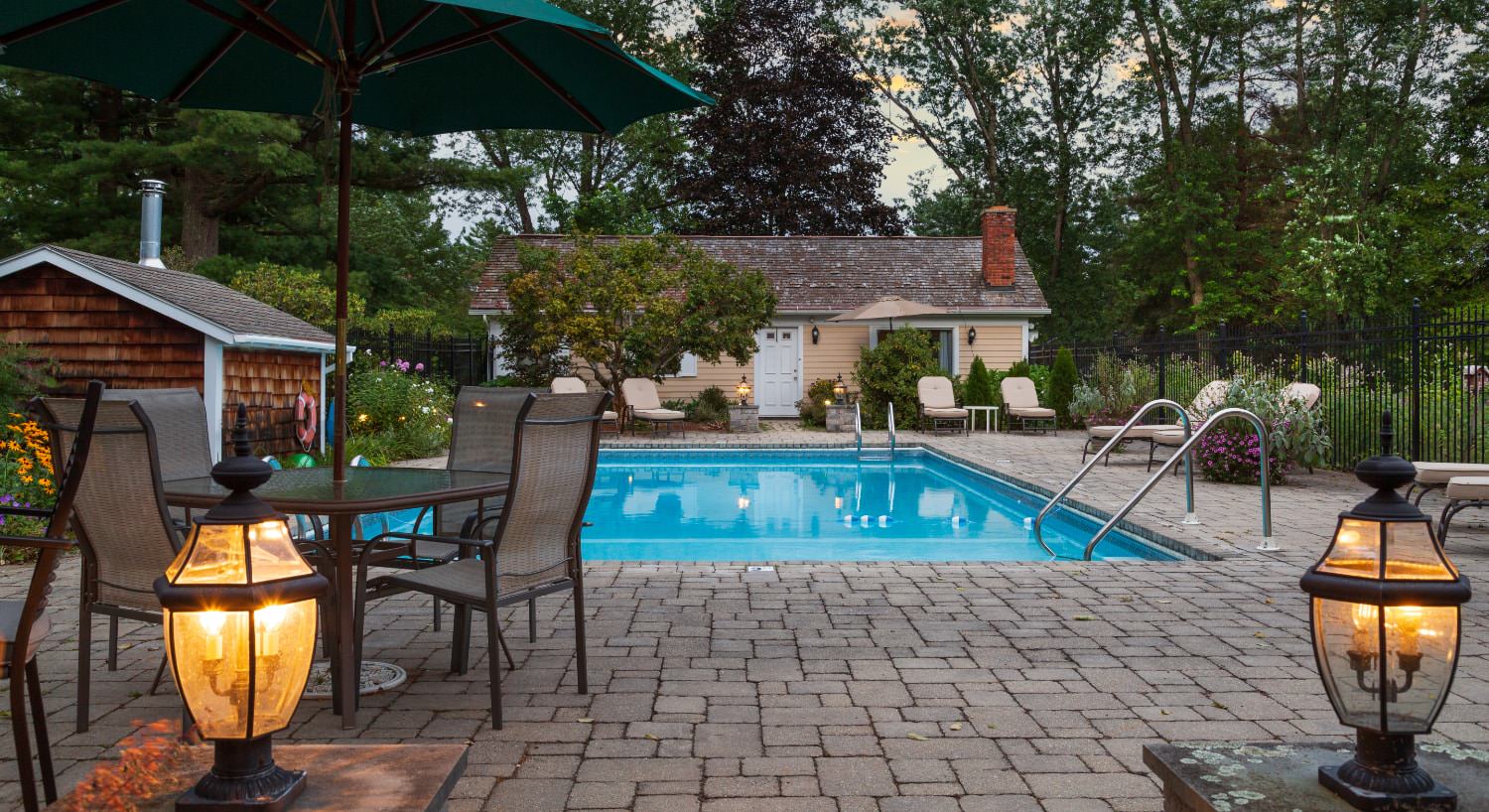 Pool area surrounded by brick patio with patio table and chairs, green umbrella, black wrought-iron fencing, large treen trees, and a lot of bushes and shrubs