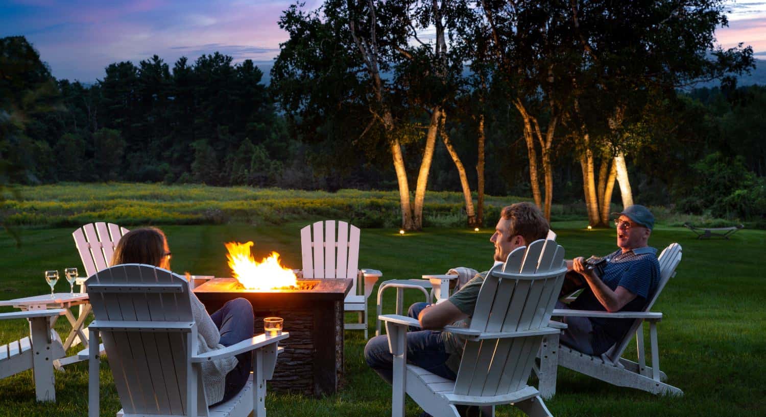 Lighted firepit surrounded by white adirondack chairs on green grass with lighted trees in the background at dusk