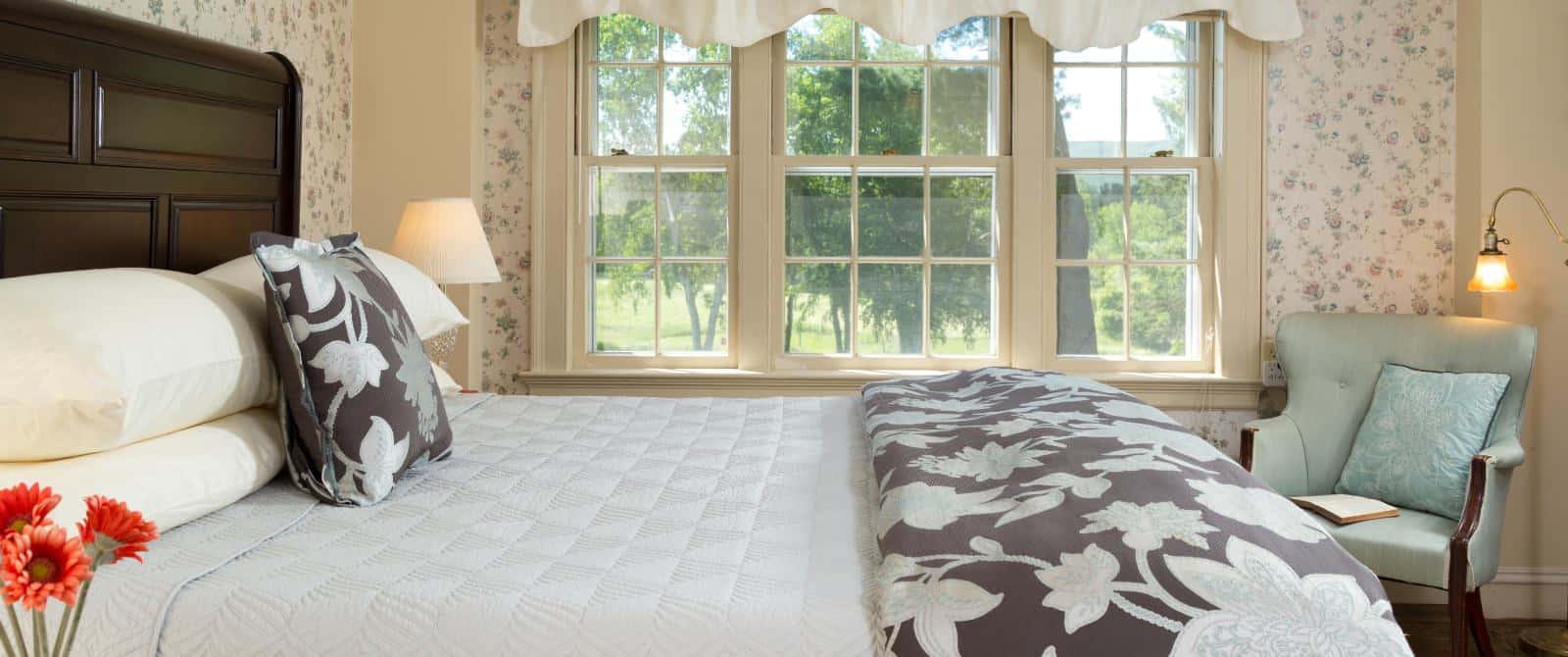 Bedroom with floral wallpaper, hardwood flooring, wooden bed, multiple hues of blue bedding, and light blue upholstered wooden chair