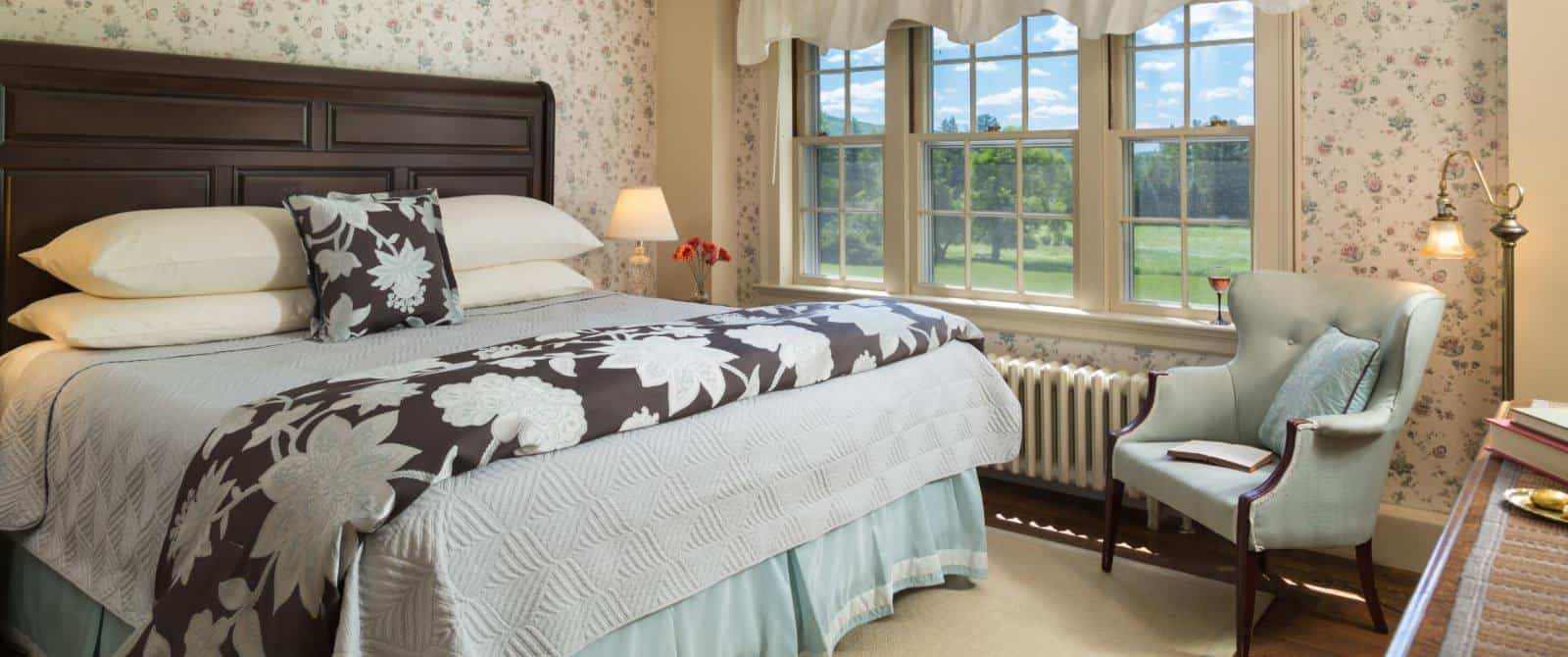 Bedroom with floral wallpaper, hardwood flooring, wooden bed, multiple hues of blue bedding, and light blue upholstered wooden chair