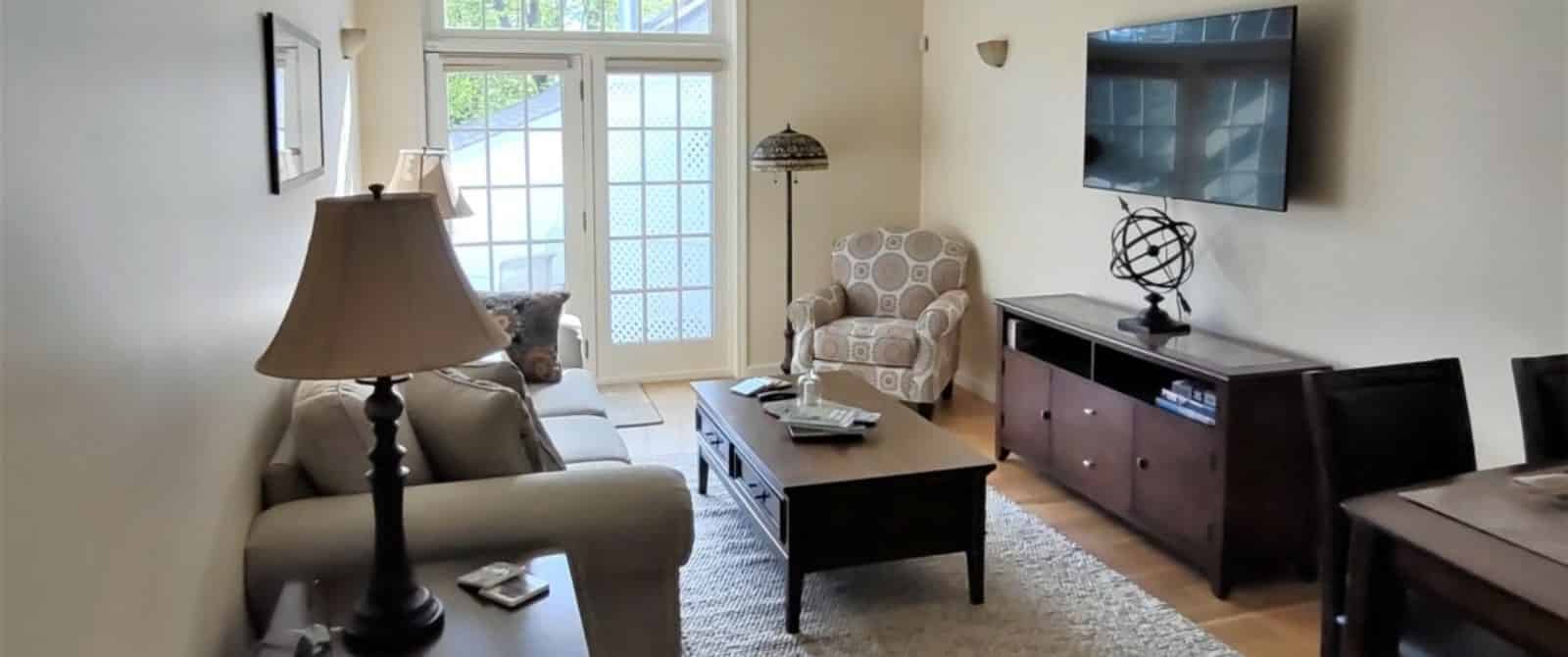 Large gathering area with light colored walls, hardwood flooring, upholstered sofa and chair, wooden coffee table, and dining area