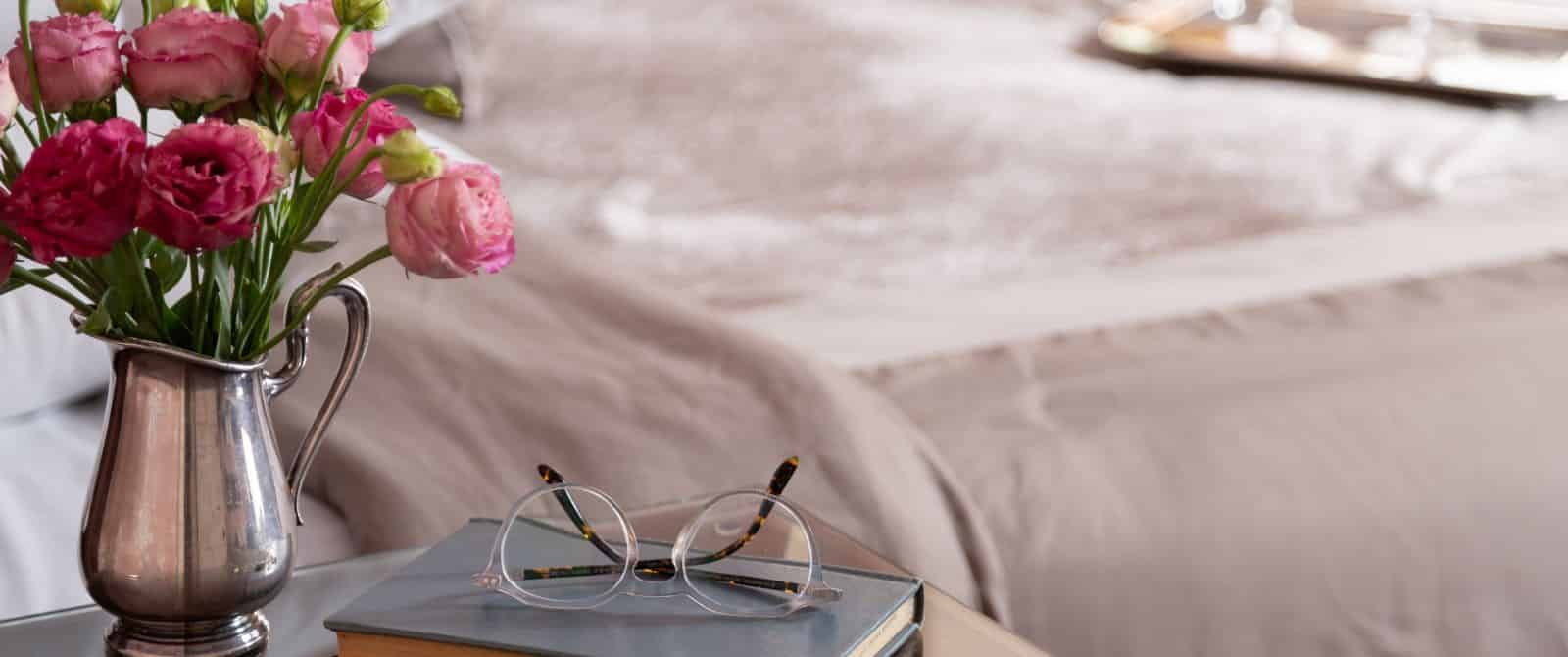 Close up view of wooden nightstand with book, vase with pink roses, and reading glasses