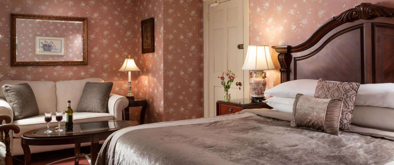 Bedroom with dark floral wallpaper, red carpeting, dark wooden headboard, gray bedding, and sitting area with upholstered sofa and dark wooden coffee table