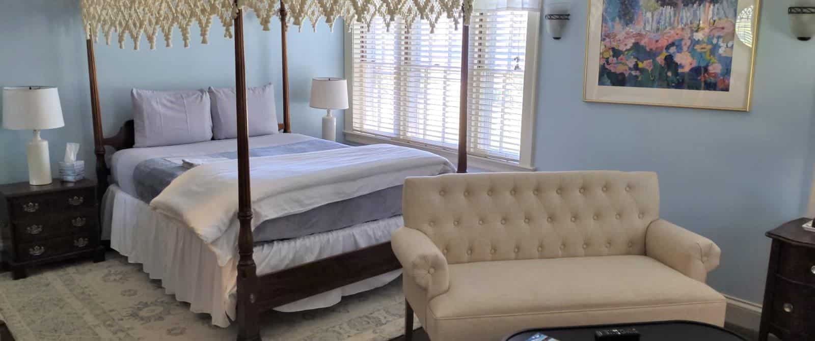 Bedroom with blue walls, white trim, hardwood flooring, dark wooden four-poster bed, lavender bedding, fireplace, and sitting area with upholstered loveseat and wooden coffee table