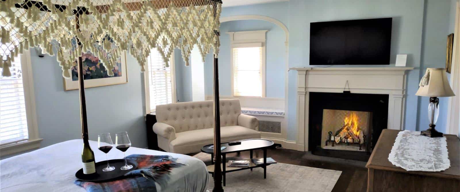 Bedroom with blue walls, white trim, hardwood flooring, dark wooden four-poster bed, lavender bedding, fireplace, wall-mounted TV, and sitting area with upholstered loveseat and wooden coffee table