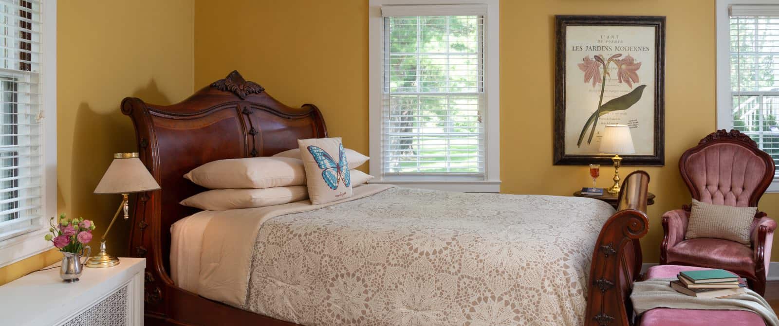 Bedroom with yellow walls, white trim, hardwood flooring, wooden sleigh bed, pink upholstered antique chair, and multiple windows