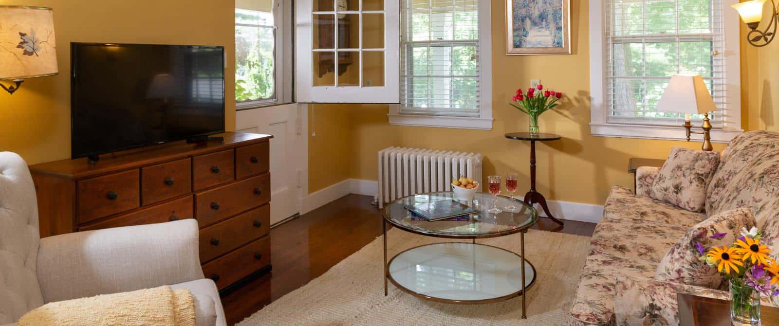 Gathering room with yellow walls, hardwood flooring, upholstered sofa and chair, glass coffee table, flat-screen TV, and views of the outside