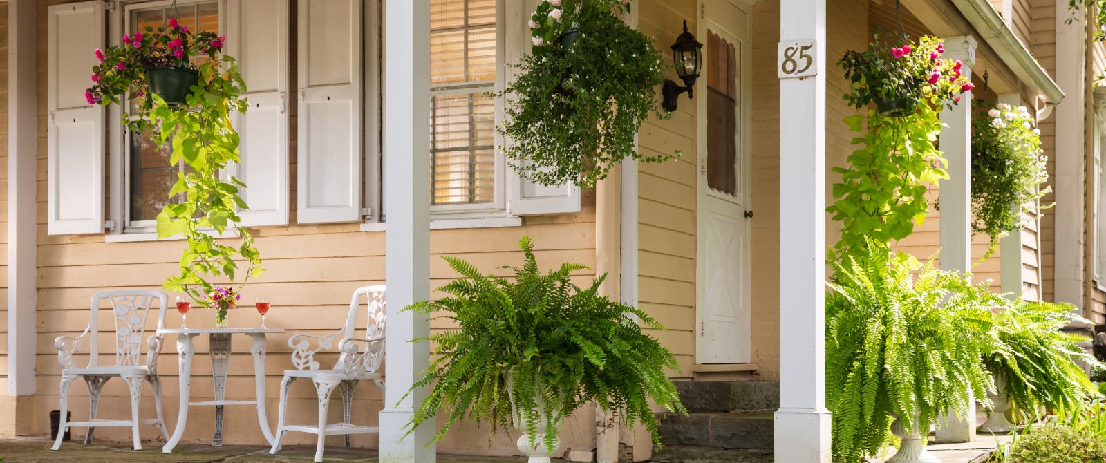 Exterior view of the property painted cream with white trim with white wrought iron table and chairs on the front porch and hanging flower baskets