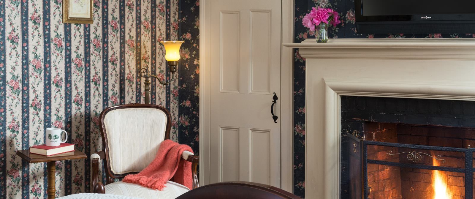 Bedroom with floral and striped wallpaper, wooden bed, fireplace, and antique wooden chair