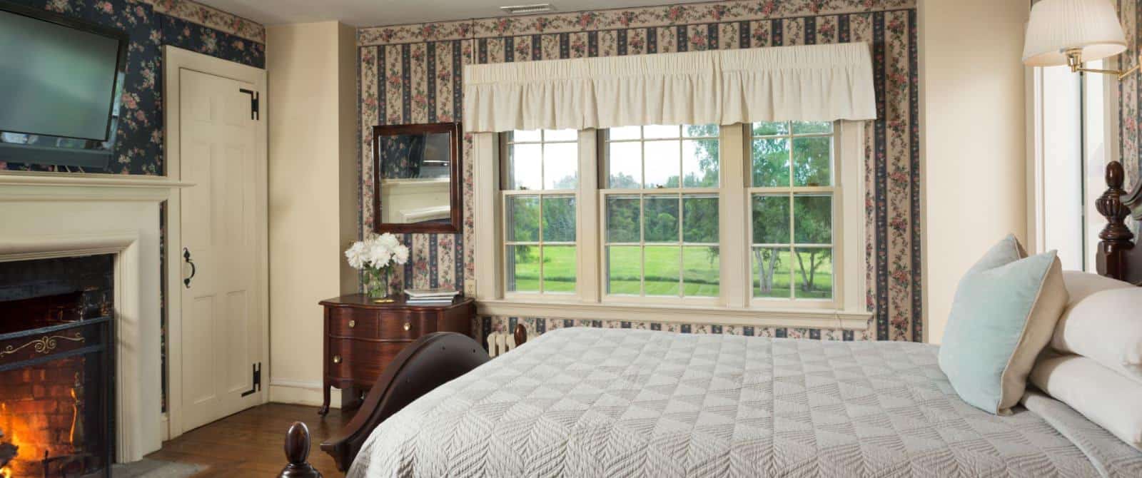 Bedroom with floral and striped wallpaper, hardwood flooring, wooden bed, light gray bedding, fireplace, and large window