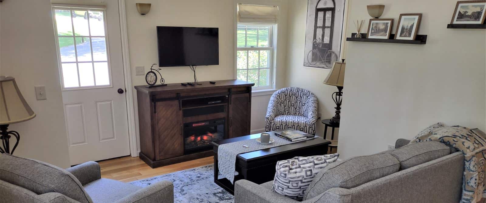 Gathering room with light colored walls, hardwood flooring, upholstered coach and chair, wooden coffee table, and wall-mounted TV