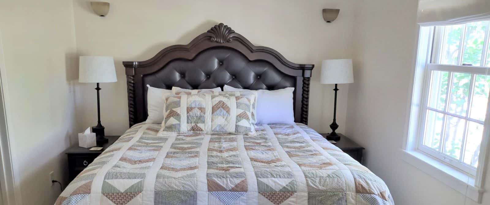 Bedroom with white walls, dark wood and leather upholstered headboard, multicolored bedding, two nightstands, and two lamps