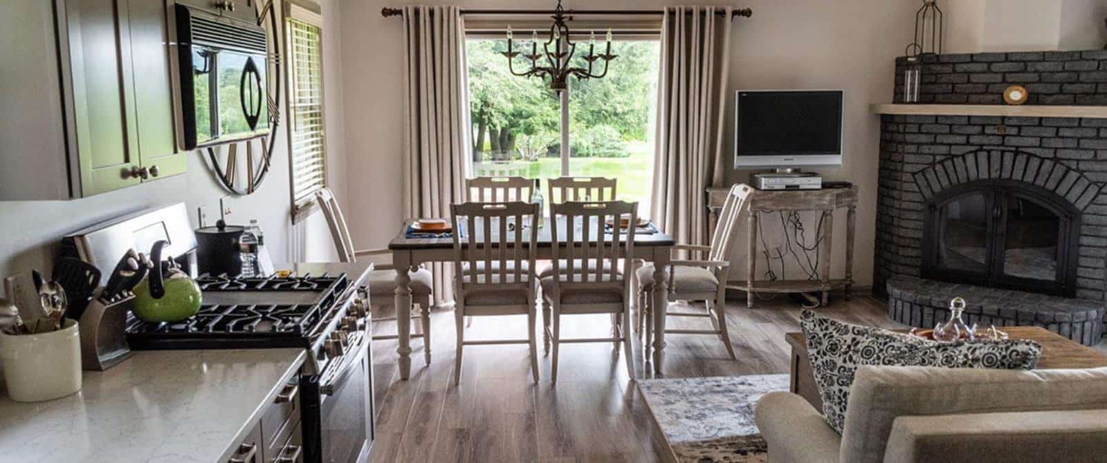 Large gathering area with light colored walls, hardwood flooring, upholstered sofa and chair, wooden coffee table, fireplace, kitchen, and dining area