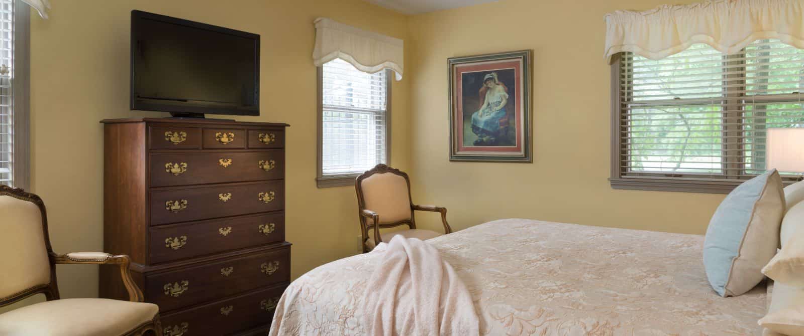 Bedroom with yellow walls, gray trim, peach bedding, wooden dresser, antique chairs, and flat-screen TV