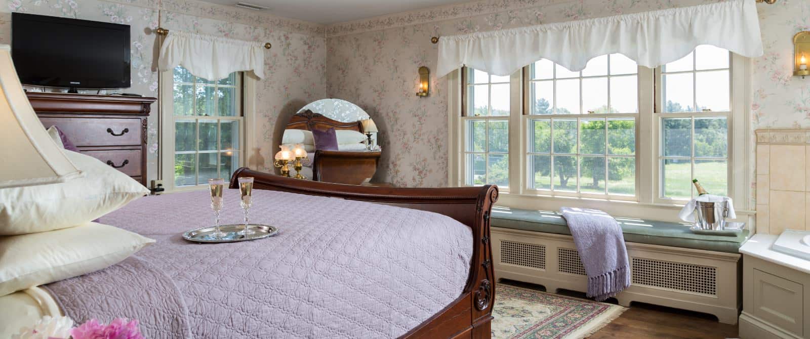 Bedroom with floral wallpaper, hardwood flooring, wooden sleigh bed, lavender bedding, wooden dresser, and jetted tub