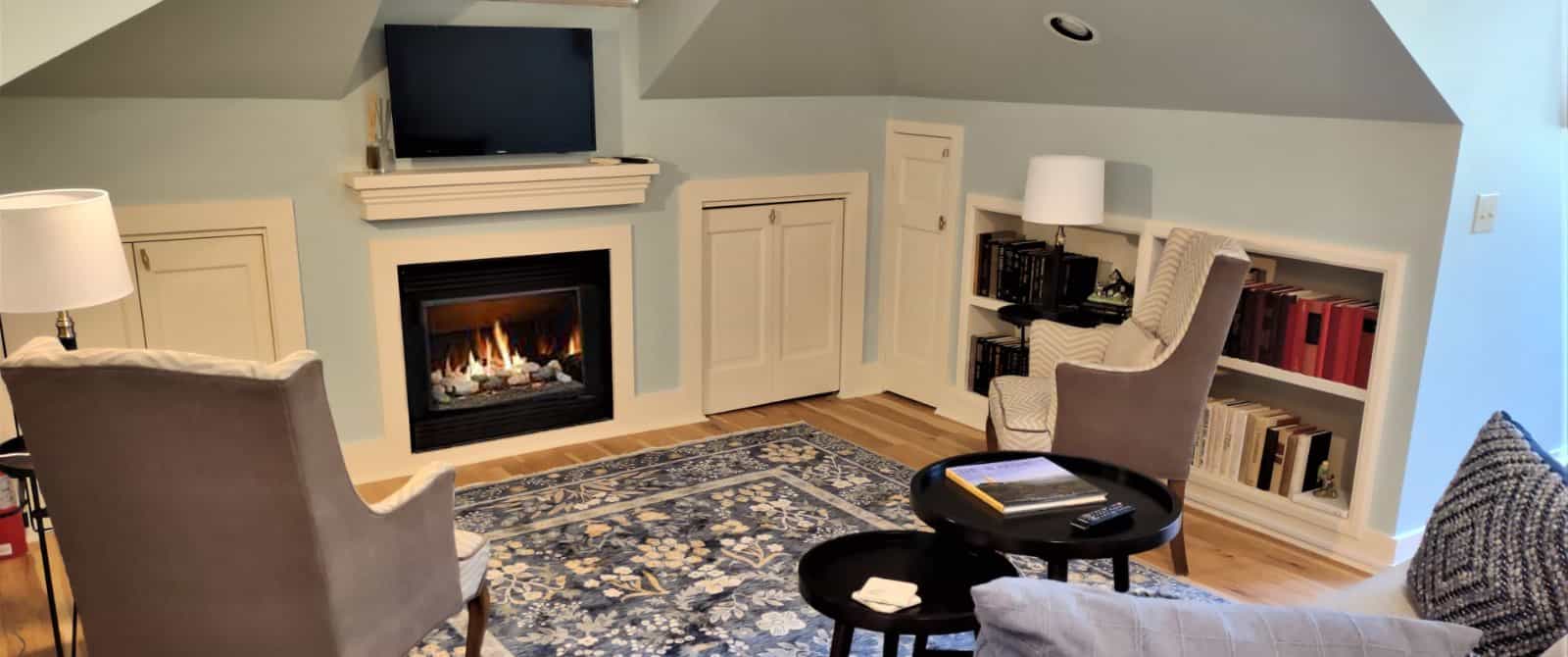 Gathering area with light blue walls, white trim, hardwood flooring, upholstered sofa, upholstered wing chairs, circular tables, fireplace, and wall-mounted TV