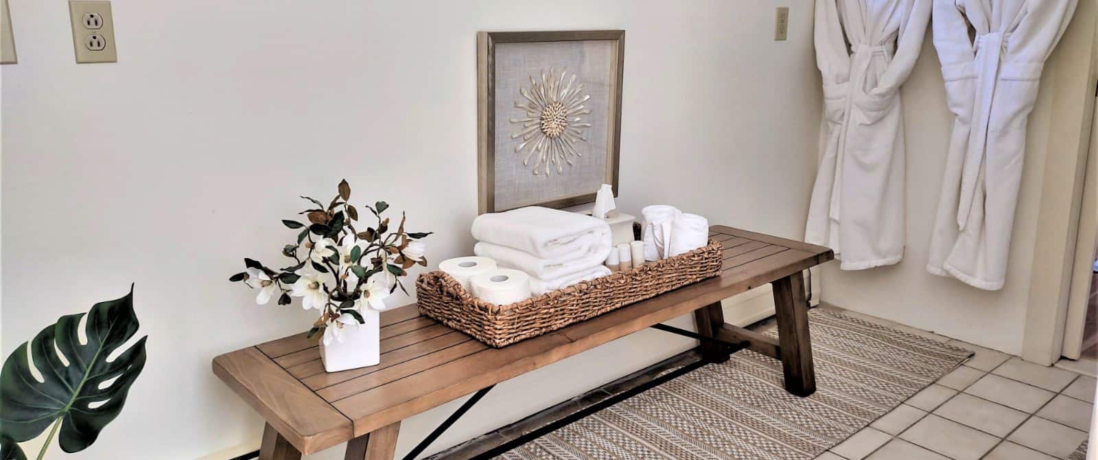 Bathroom with a long wooden bench with a basket filled with toilet paper, towels, and toiletries