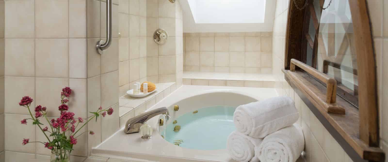 Large jetted tub full of water surrounded by light tan tile, three rolled white towels, and vase of pink flowers